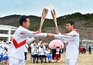 Ryoji Sakuma (right) receiving the Olympic frame at a "torch kiss" point and fist-bumping the previous torchbearer (left). His running course was in Katsurao Primary School, which is the old school for Ryoji, his father, and his grandfather. (in Katsurao Village, Fukushima Prefecture)